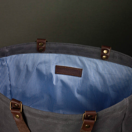 Gray tote bag with blue interior lining and brown leather straps on a dark background