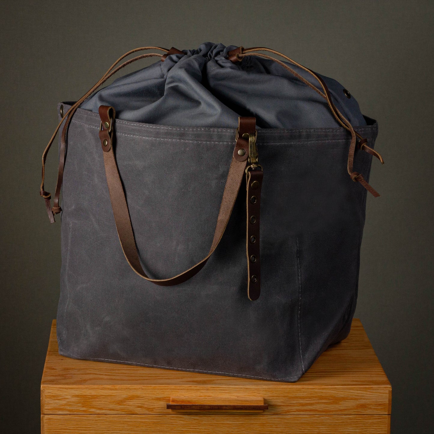 Gray drawstring bag with brown leather straps on a wooden surface against a dark background