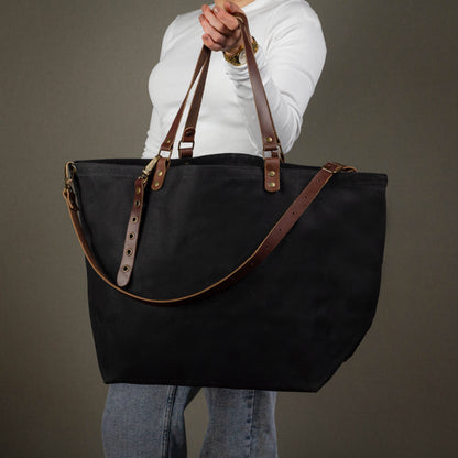 Person holding a black tote bag with brown straps against a gray background