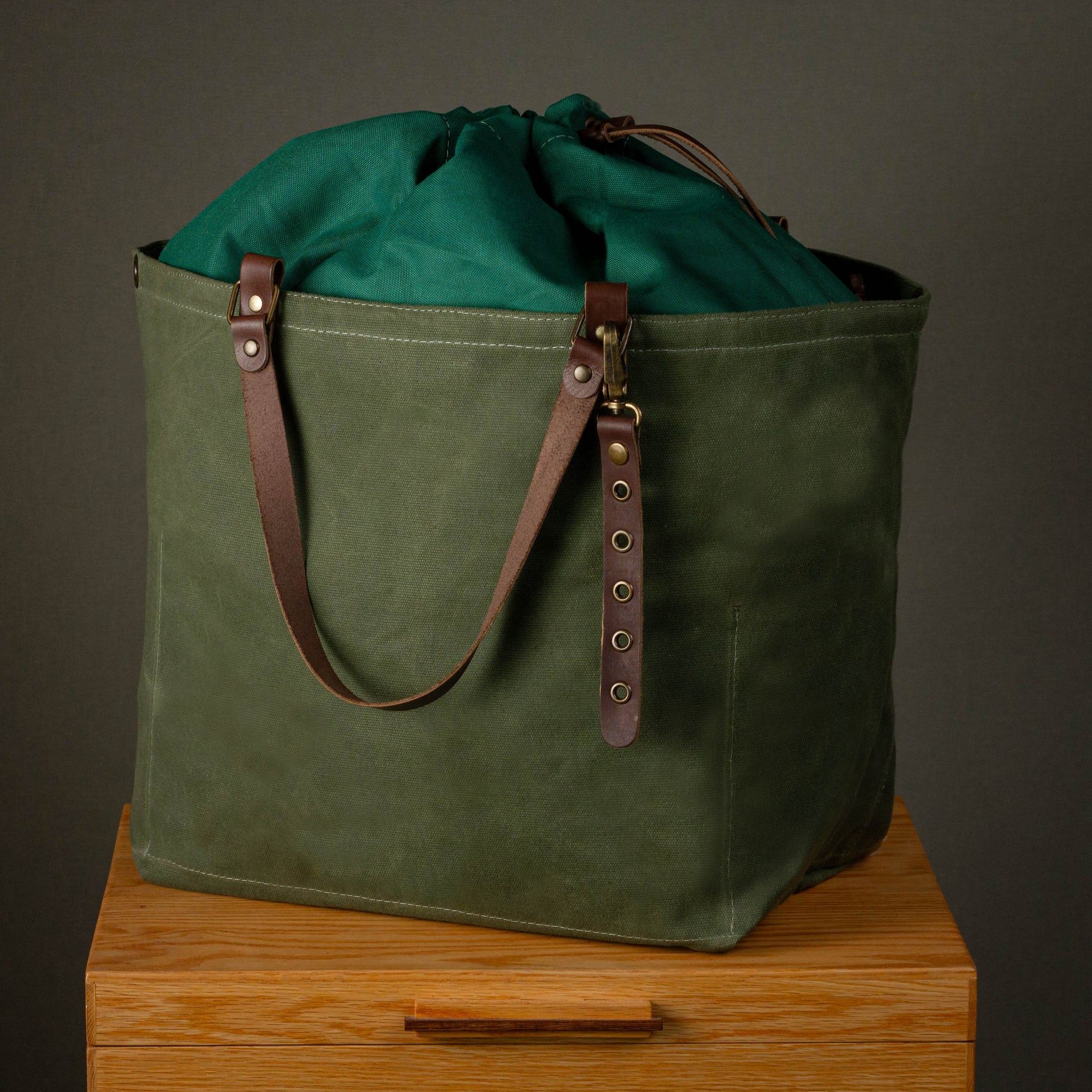 Green waxed canvas bag with leather straps on a wooden surface against a dark background