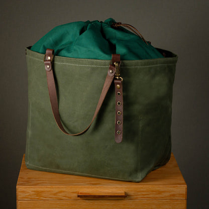 Green waxed canvas bag with leather straps on a wooden surface against a dark background