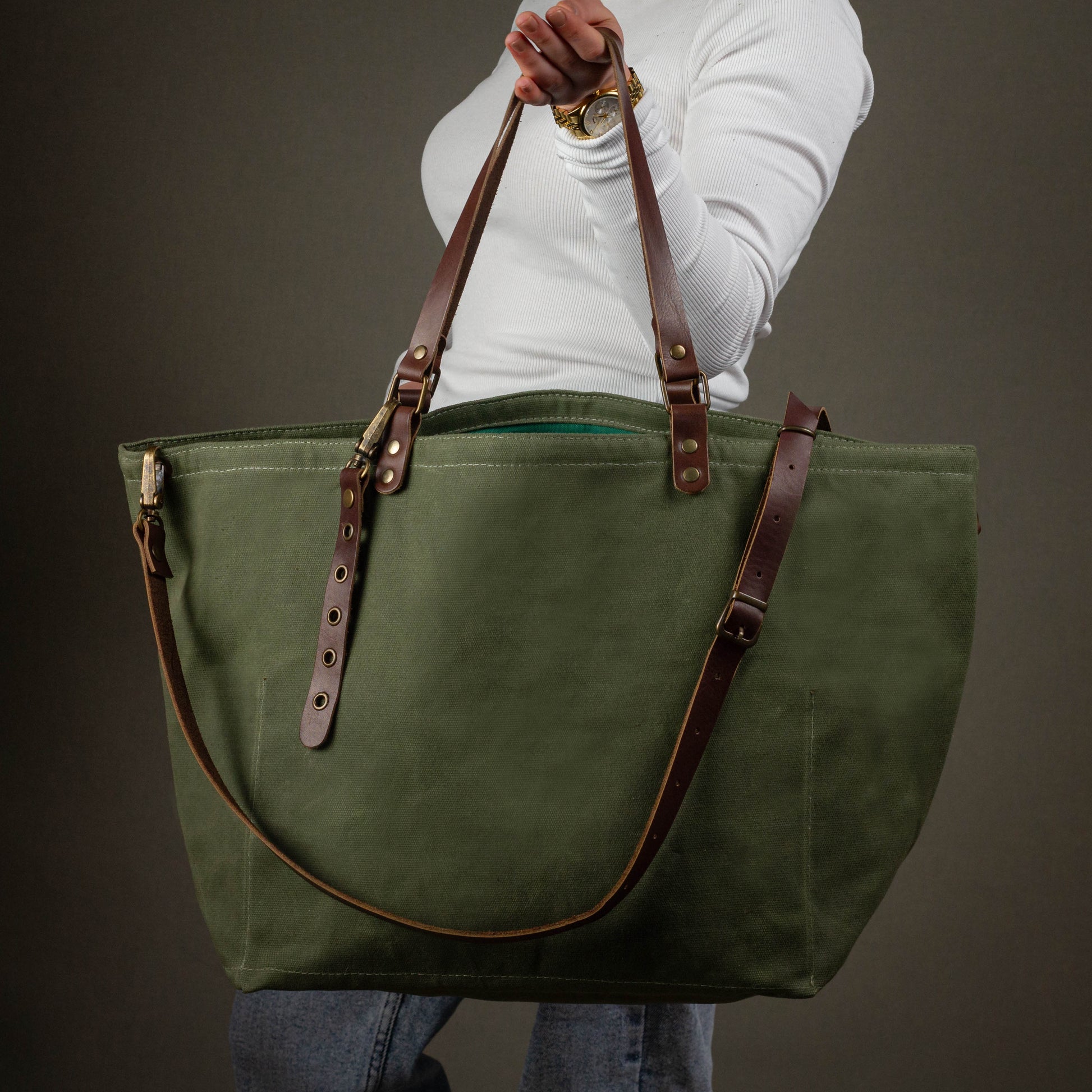 Green tote bag with brown leather straps held by a person against a gray background