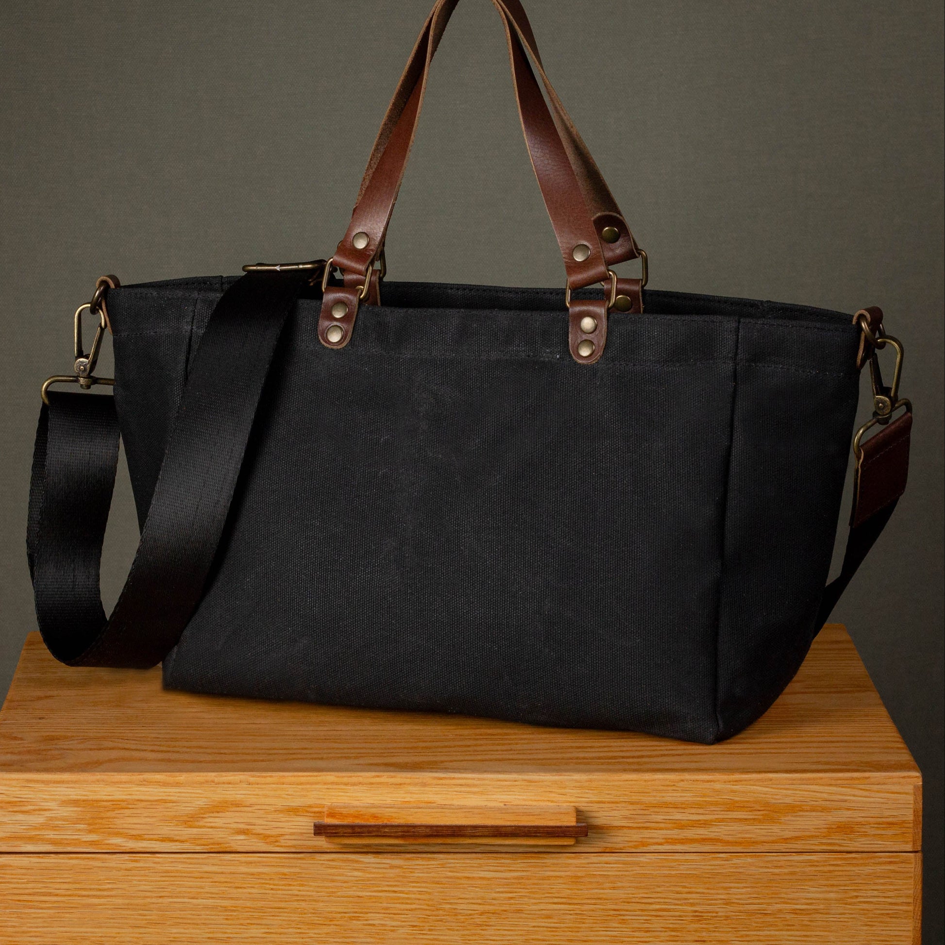 Black tote bag with brown handles and straps on a wooden surface with a dark background