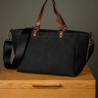 Black tote bag with brown handles and straps on a wooden surface with a dark background