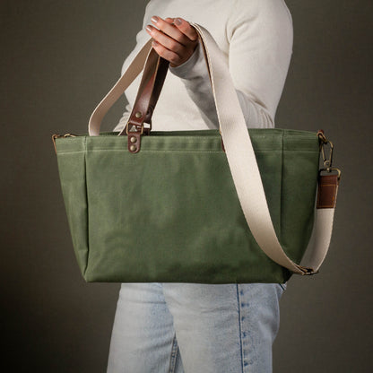 Green tote bag with brown straps held by a person against a dark background