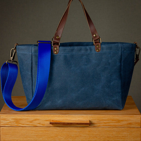 Blue tote bag with brown handles and blue strap on a wooden surface with a dark background