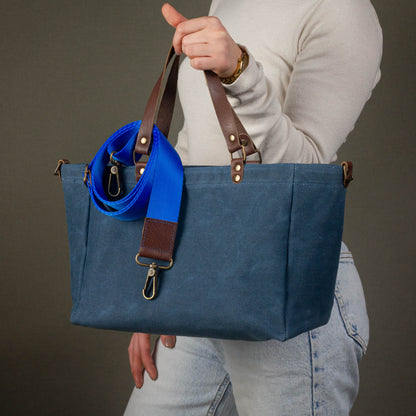 Person holding a blue tote bag with brown straps against a gray background