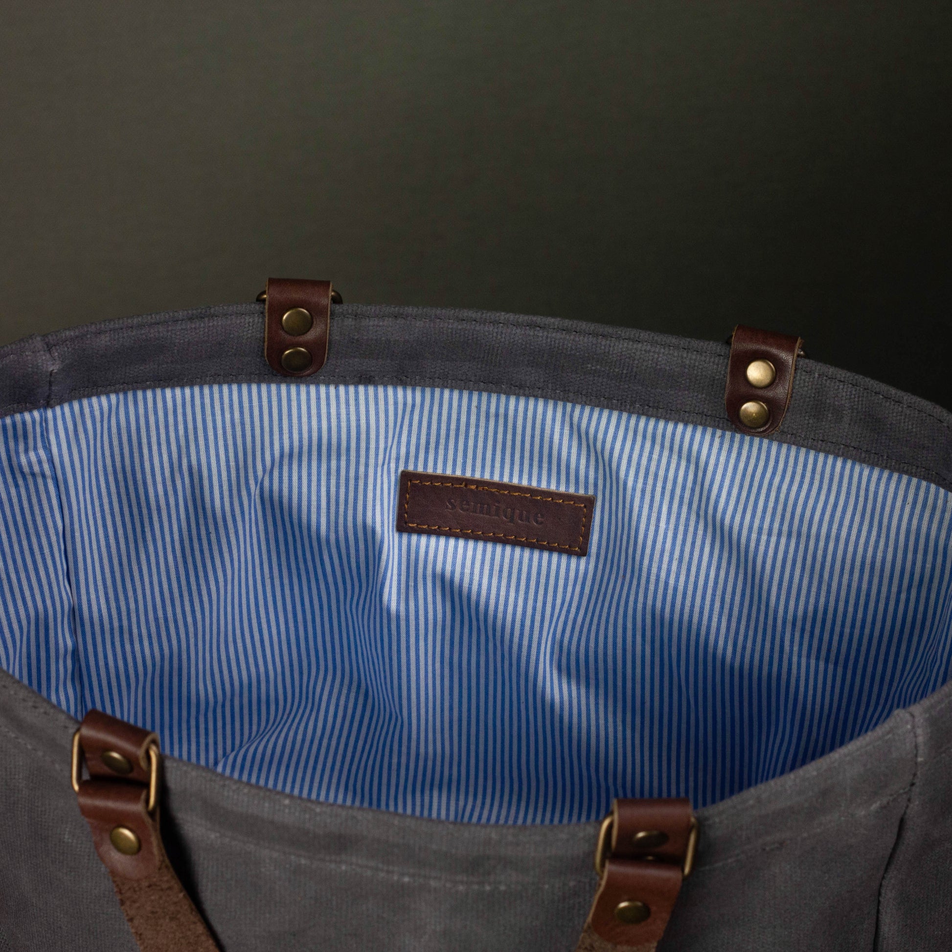 Gray tote bag with blue interior lining and brown leather straps on a dark background