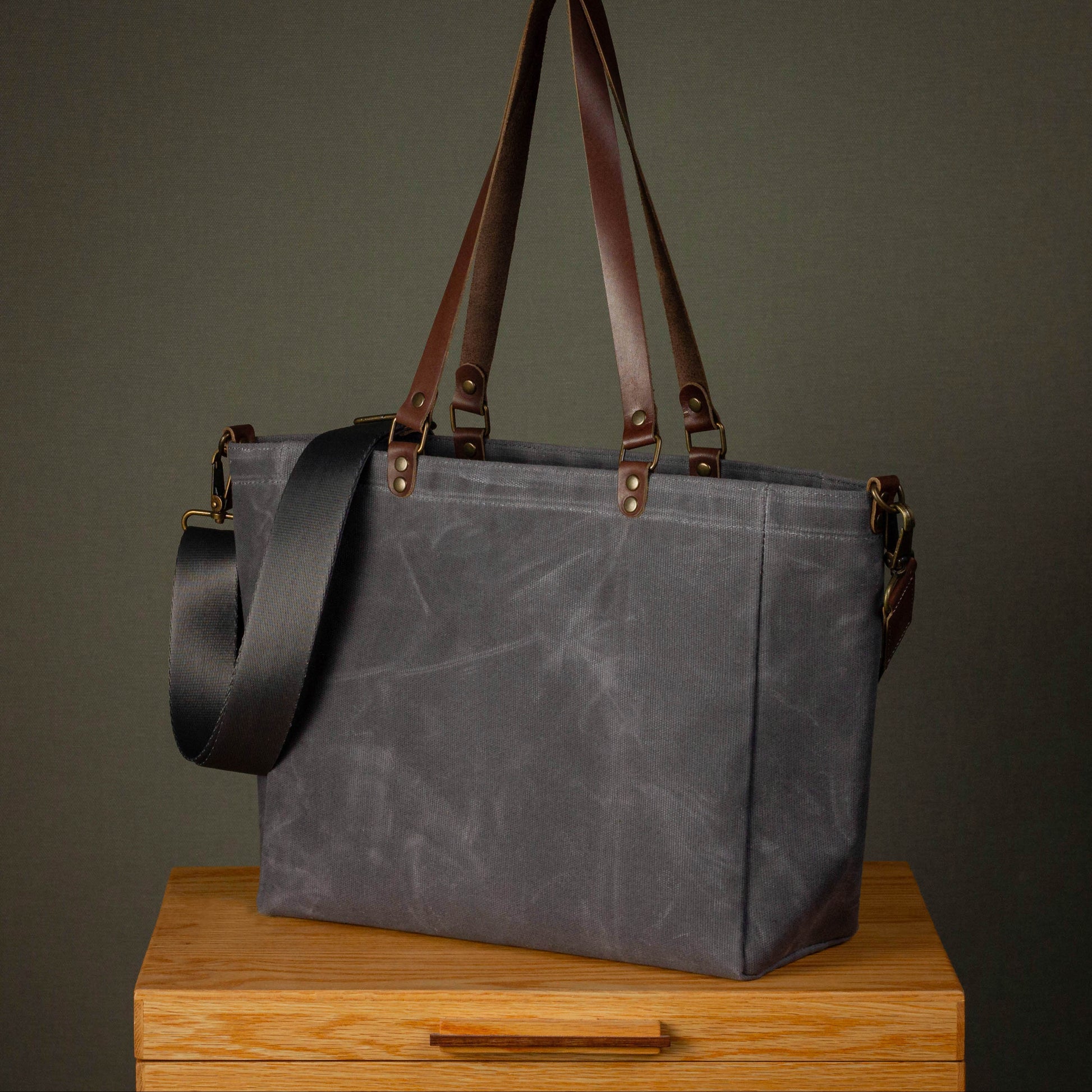 Gray tote bag with brown handles and strap on a wooden surface against a dark background