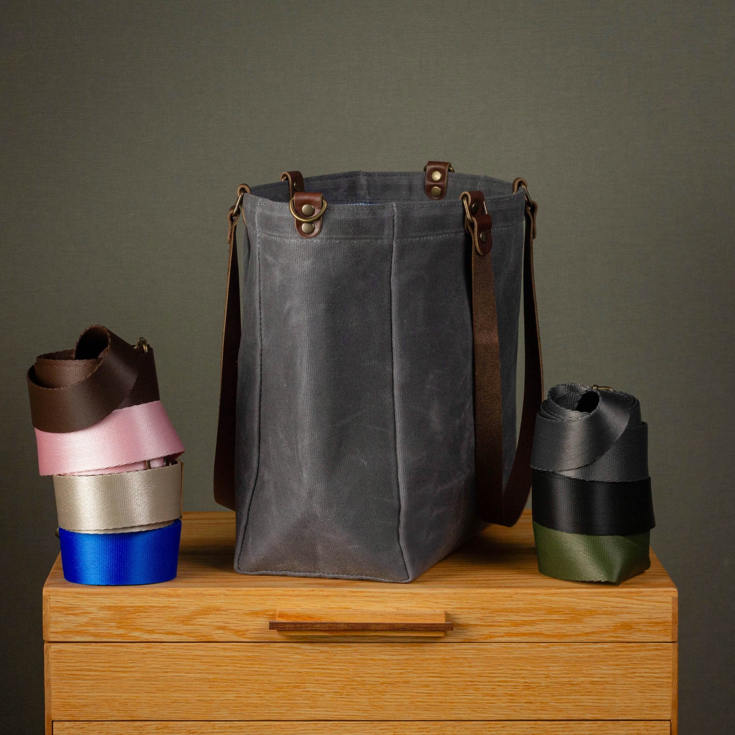 Gray waxed canvas bag with brown leather straps on a wooden surface against a gray background