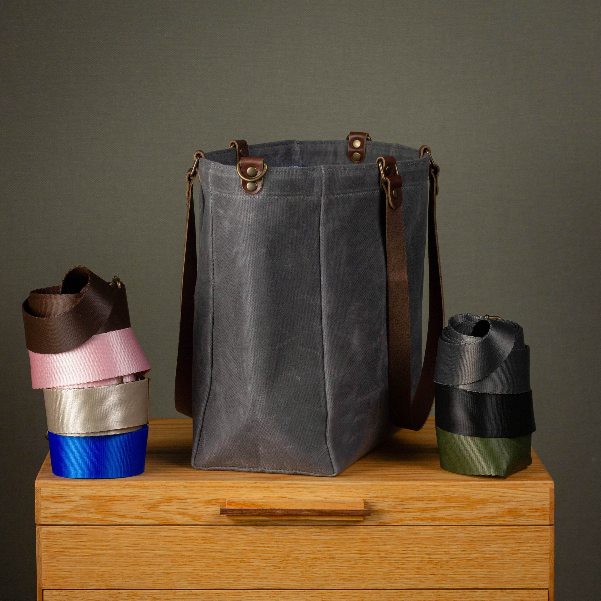 Gray waxed canvas bag with brown leather straps on a wooden surface against a gray background