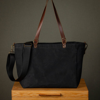 Black tote bag with brown straps on a wooden surface against a dark background
