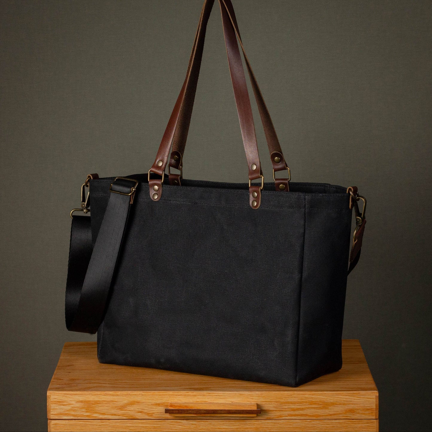 Black tote bag with brown straps on a wooden surface against a dark background