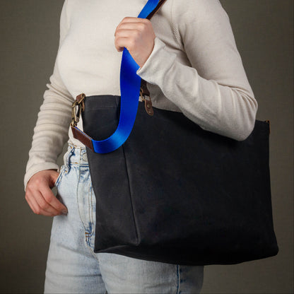 Person holding a black tote bag with a blue strap against a gray background