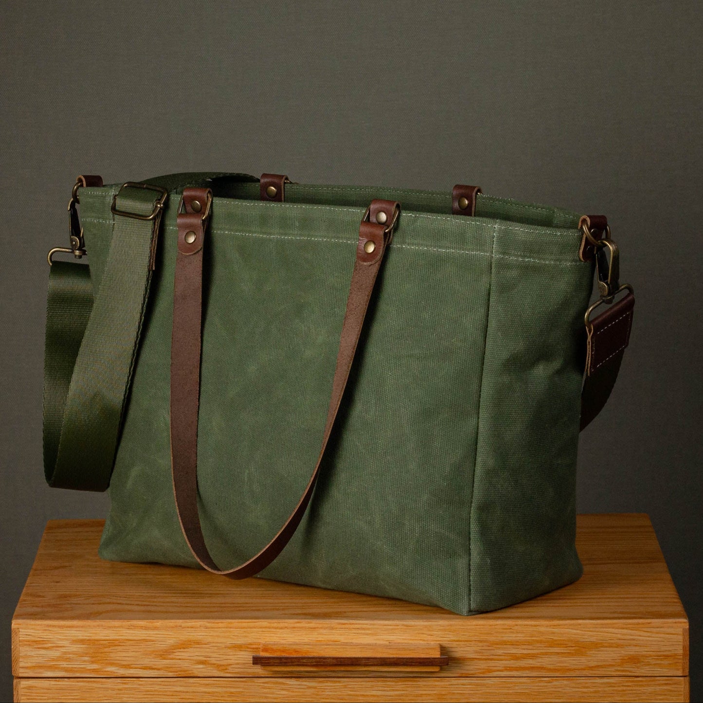 Green canvas bag with brown straps on a wooden surface against a gray background